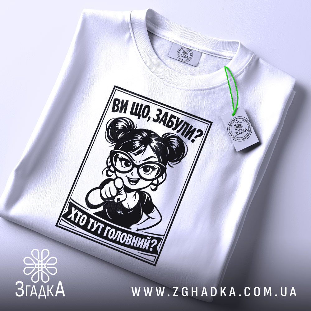 Футболка хто тут головний. Біла футболка, складена на поверхні, з принтом чорного кольору. На футболці видно персонажа з окулярами, який вказує пальцем. На футболці є етикетка з зеленим шнурком. Фон світло-сірий.