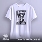 Футболка хто тут головний. Біла футболка, підвішена на вішалці, з принтом чорного кольору, що зображує персонажа з окулярами. Фон світло-сірий, деталі принта добре проглядаються.