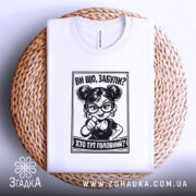 Футболка хто тут головний. Біла футболка, складена на плетеній основі, з принтом чорного кольору, що зображує персонажа з окулярами. Фон білий, деталі принта чітко видимі.