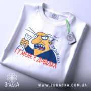 Футболка художник маю довідку. Біла футболка, складена на поверхні, з принтом на передній частині. Принт зображає персонажа з жовтим обличчям, який тримає аркуш паперу, з написом 'Я художник! І у мене є довідка'. На футболці є етикетка з логотипом бренду.