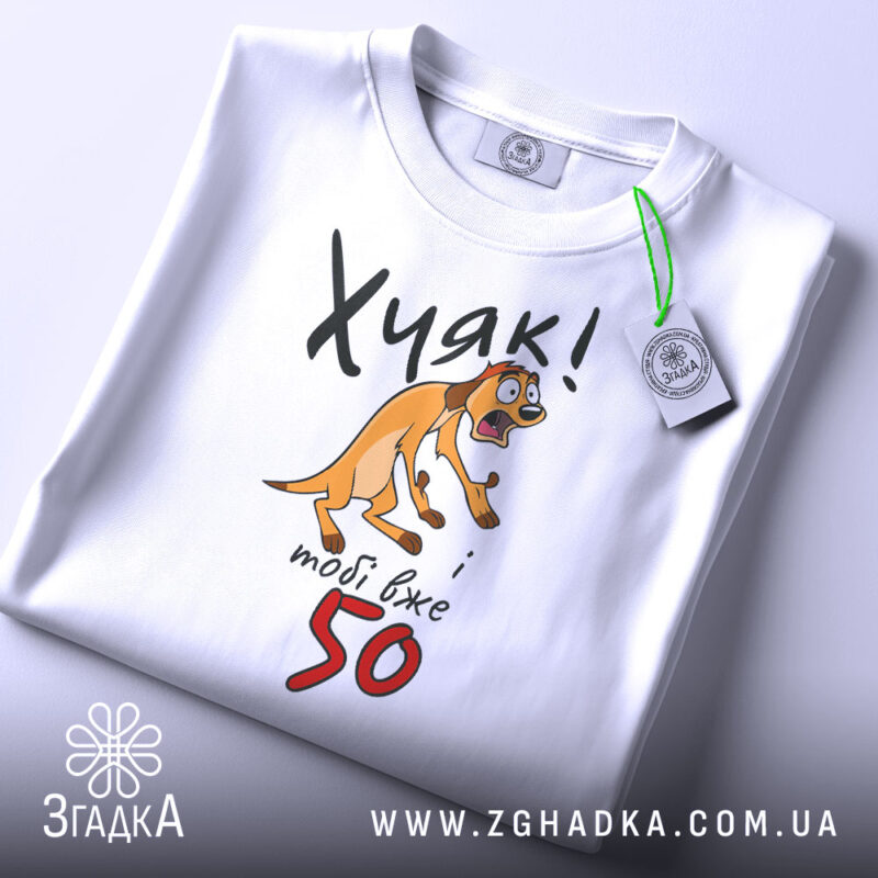 Футболка Хуяк тобі 50, біла, складена на поверхні, з видимим етикеткою, фон світло-сірий, принт з жовтим собакою, що виглядає здивованою, текст чорний і червоний.