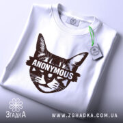 Футболка кіт анонімус білого кольору, складена на поверхні, фон світло-сірий, принт зображає кота з написом 'ANONYMOUS' у коричневому кольорі, на футболці є етикетка з зеленим шнурком.
