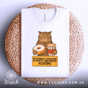 Футболка кіт і кава, біла, зображена зверху на плетеному кошику, фон білий, принт з котом, який тримає пончик і чашку кави, текст на жовтому фоні: 'половину проблем в житті вирішує кофе'.