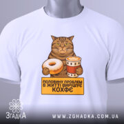 Футболка кіт і кава, біла, зображена зверху, на столі, фон світло-сірий, принт з котом, який тримає пончик і чашку кави, текст на жовтому фоні: 'половину проблем в житті вирішує кофе', на футболці є етикетка.