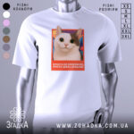 Футболка кіт білого кольору, зображена на манекені, фон світло-сірий, принт з великим котом з великими очима на кольоровому фоні з текстом.