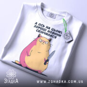 Футболка кіт погані новини, біла, складена на поверхні, фон світлий, принт з котом з ножем, з рожевими і жовтими відтінками, з етикеткою на рукаві.