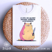 Футболка кіт погані новини, біла, складена на плетеній кошику, фон світлий, принт з котом з ножем, з рожевими і жовтими відтінками.