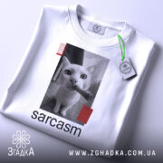 Футболка кіт сарказм, біла, складена на столі, фон світло-сірий, принт з чорним котом, який тримає сигару, з червоними смужками, з етикеткою.