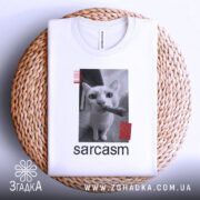 Футболка кіт сарказм, біла, складена на плетеному кошику, фон світло-сірий, принт з чорним котом, який тримає сигару, з червоними смужками.