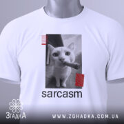 Футболка кіт сарказм, біла, складена на столі, фон світло-сірий, принт з чорним котом, який тримає сигару, з червоними смужками, з етикеткою.