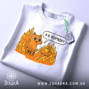 Футболка кіт це нормально. Біла футболка, складена на столі, з принтом оранжевого кота на стільці серед вогню, з текстом 'Я в порядку'. На футболці є етикетка з зеленим шнурком. Фон світло-сірий.