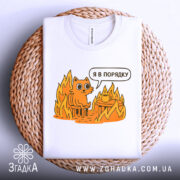 Футболка кіт це нормально. Біла футболка, складена на плетеній основі, з принтом оранжевого кота на стільці серед вогню, з текстом 'Я в порядку'. Фон світло-сірий, деталі принта яскраві.