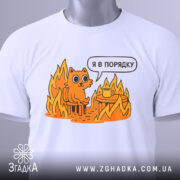 Футболка кіт це нормально. Біла футболка, складена на столі, з принтом оранжевого кота на стільці серед вогню, з текстом 'Я в порядку'. На футболці є етикетка з зеленим шнурком. Фон світло-сірий.
