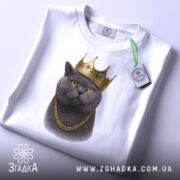 Футболка кіт у короні, біла, складена на поверхні, фон світло-сірий, принт зображає кота в золотій короні та ланцюгу, з етикеткою з зеленим шнурком.