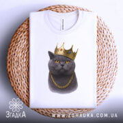Футболка кіт у короні, біла, складена на плетеній кошику, фон білий, принт зображає кота в золотій короні та ланцюгу, з чіткими деталями.