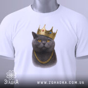 Футболка кіт у короні, біла, складена на поверхні, фон світло-сірий, принт зображає кота в золотій короні та ланцюгу, з етикеткою з зеленим шнурком.