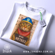 Футболка кіт у розшуку, біла, складена на столі, фон світло-сірий, принт з котом у блакитній шапці та червоному бандану, на фоні ліній для зросту з написом ID NO. 987602, з етикеткою на шнурку.