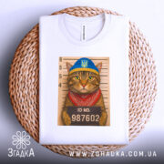 Футболка кіт у розшуку, біла, складена на круглому плетеному кошику, фон світло-сірий, принт з котом у блакитній шапці та червоному бандану, на фоні ліній для зросту з написом ID NO. 987602.