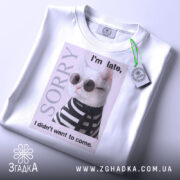 Футболка кіт у стилі білого кольору, зображена на столі, фон світло-сірий, принт з котом у сонцезахисних окулярах та смугастому светрі, текст на футболці говорить 'I'm late, I didn't want to come.', на футболці є етикетка з зеленим шнурком.