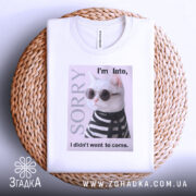 Футболка кіт у стилі білого кольору, зображена на плетеному кошику, фон світлий, принт з котом у сонцезахисних окулярах та смугастому светрі, текст на футболці говорить 'I'm late, I didn't want to come.'