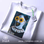 Футболка клоун жахів, біла, складена на столі, фон світло-сірий, принт з клоуном у соняшникових окулярах, прикріплена етикетка з логотипом.