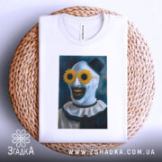 Футболка клоун жахів, біла, розташована на плетеній основі, фон світло-сірий, принт з клоуном у соняшникових окулярах, видно етикетку на комірці.