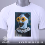 Футболка клоун жахів, біла, складена на столі, фон світло-сірий, принт з клоуном у соняшникових окулярах, прикріплена етикетка з логотипом.