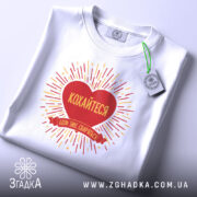 Футболка кохайтеся коли сваряться, біла, складена на столі, фон світло-сірий, принт червоного серця з жовтими променями та написом, на комірці видна етикетка.