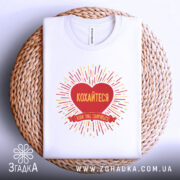 Футболка кохайтеся коли сваряться, біла, складена на плетеній кошику, фон білий, принт червоного серця з жовтими променями та написом.