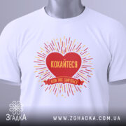 Футболка кохайтеся коли сваряться, біла, складена на столі, фон світло-сірий, принт червоного серця з жовтими променями та написом, на комірці видна етикетка.