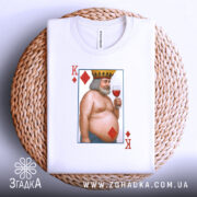 Футболка король черви, біла, складена на плетеній кошику, фон світло-сірий, принт з ілюстрацією короля червів, який тримає келих з вином, з короною на голові, з червоними ромбами.