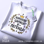 Футболка королева я легенда, біла, зображена в складеному вигляді, на світлому фоні, принт з текстом "Королева? Цариця? Я – легенда!" яскравий, з короною та сердечками, прикріплений етикеткою зеленого кольору.