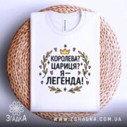 Футболка королева я легенда, біла, зображена в складеному вигляді, на плетеній основі, принт з текстом "Королева? Цариця? Я – легенда!" яскравий, з короною та сердечками, виглядає елегантно.
