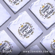 Футболка королева я легенда, біла, зображена з фронтального ракурсу, на нейтральному фоні, принт з текстом "Королева? Цариця? Я – легенда!" оформлений у кольорах жовтий, чорний, рожевий та зелений, оточений декоративними елементами.