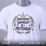 Футболка королева я легенда, біла, зображена в складеному вигляді, на світлому фоні, принт з текстом "Королева? Цариця? Я – легенда!" яскравий, з короною та сердечками, прикріплений етикеткою зеленого кольору.