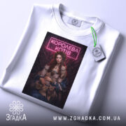 Футболка королева котів, біла, лежить на столі, фон світло-сірий, принт з дівчиною в блакитній сукні, яка тримає котів, з неоновим написом 'КОРОЛЕВА КОТІВ', на футболці є етикетка з зеленим шнурком.