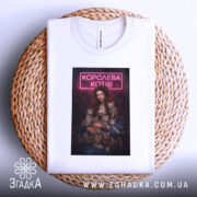 Футболка королева котів, біла, лежить на плетеній кошику, фон світло-сірий, принт з дівчиною в блакитній сукні, яка тримає котів, з неоновим написом 'КОРОЛЕВА КОТІВ'.
