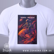 Футболка космос мій океан, біла, складена на поверхні, фон світлий, принт з космонавтом на рибі в космосі, прикріплена етикетка з зеленим шнурком.