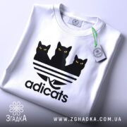 Футболка котячий стиль adicats, біла, складена на поверхні, з видимим етикеткою, принт з трьома чорними котами з жовтими очима на смугастому фоні, з легким відблиском.