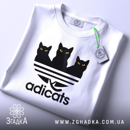 Футболка котячий стиль adicats, біла, складена на поверхні, з видимим етикеткою, принт з трьома чорними котами з жовтими очима на смугастому фоні, з легким відблиском.