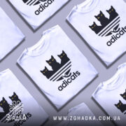 Футболка котячий стиль adicats, біла, зображена з фронтального ракурсу, на фоні світло-сірого кольору, принт з трьома чорними котами з жовтими очима на смугастому фоні.