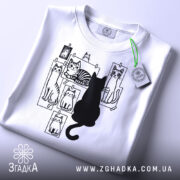 Футболка котячий світ, біла, складена на столі, фон світло-сірий, принт з чорними котами на фотографіях, один чорний кіт сидить спиною до глядача, на футболці є етикетка з зеленим шнурком.