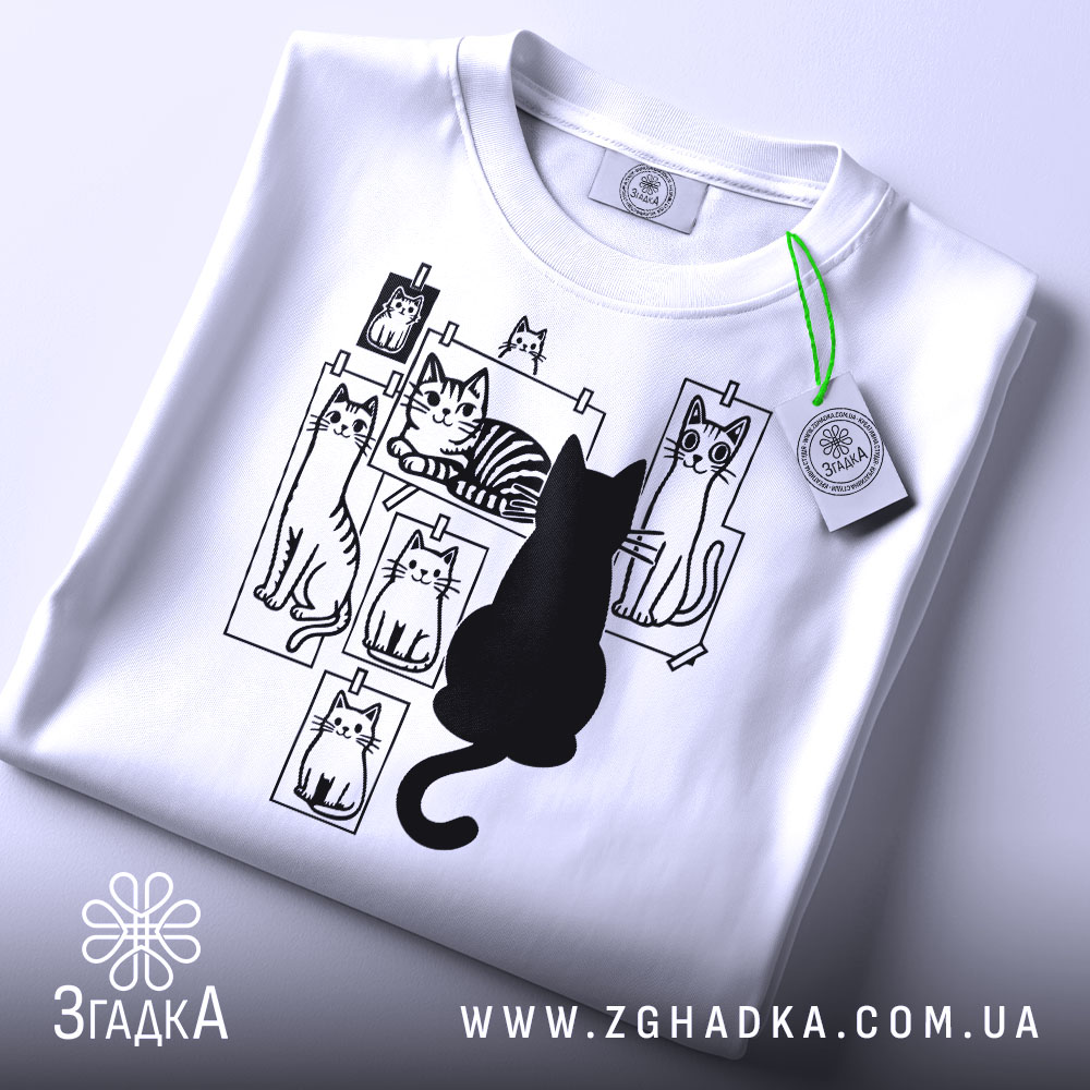 Футболка котячий світ, біла, складена на столі, фон світло-сірий, принт з чорними котами на фотографіях, один чорний кіт сидить спиною до глядача, на футболці є етикетка з зеленим шнурком.