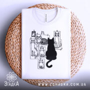 Футболка котячий світ, біла, складена на плетеній кошику, фон світло-сірий, принт з чорними котами на фотографіях, один чорний кіт сидить спиною до глядача.