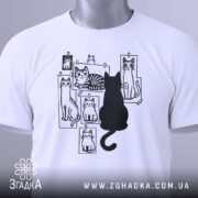 Футболка котячий світ, біла, складена на столі, фон світло-сірий, принт з чорними котами на фотографіях, один чорний кіт сидить спиною до глядача, на футболці є етикетка з зеленим шнурком.