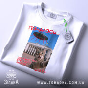 Футболка коти й прибульці, біла, складена на поверхні, фон світлий, принт з котом на фоні будівлі, текст червоним кольором, з біркою.