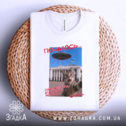 Футболка коти й прибульці, біла, складена на плетеній основі, фон світлий, принт з котом на фоні будівлі, текст червоним кольором.