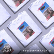 Футболка коти й прибульці, біла, зображена на манекені, фон нейтральний, принт з котом, що виглядає з-під літаючої тарілки, текст червоним кольором.