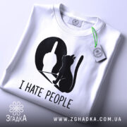 Футболка коти ненавиджу людей, біла, складена на плоскій поверхні, фон світло-сірий, принт чорного кольору з силуетом кота та жестом руки, на комірці є етикетка.