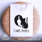 Футболка коти ненавиджу людей, біла, складена на плоскій поверхні з плетеним кошиком на фоні, принт чорного кольору з силуетом кота та жестом руки.